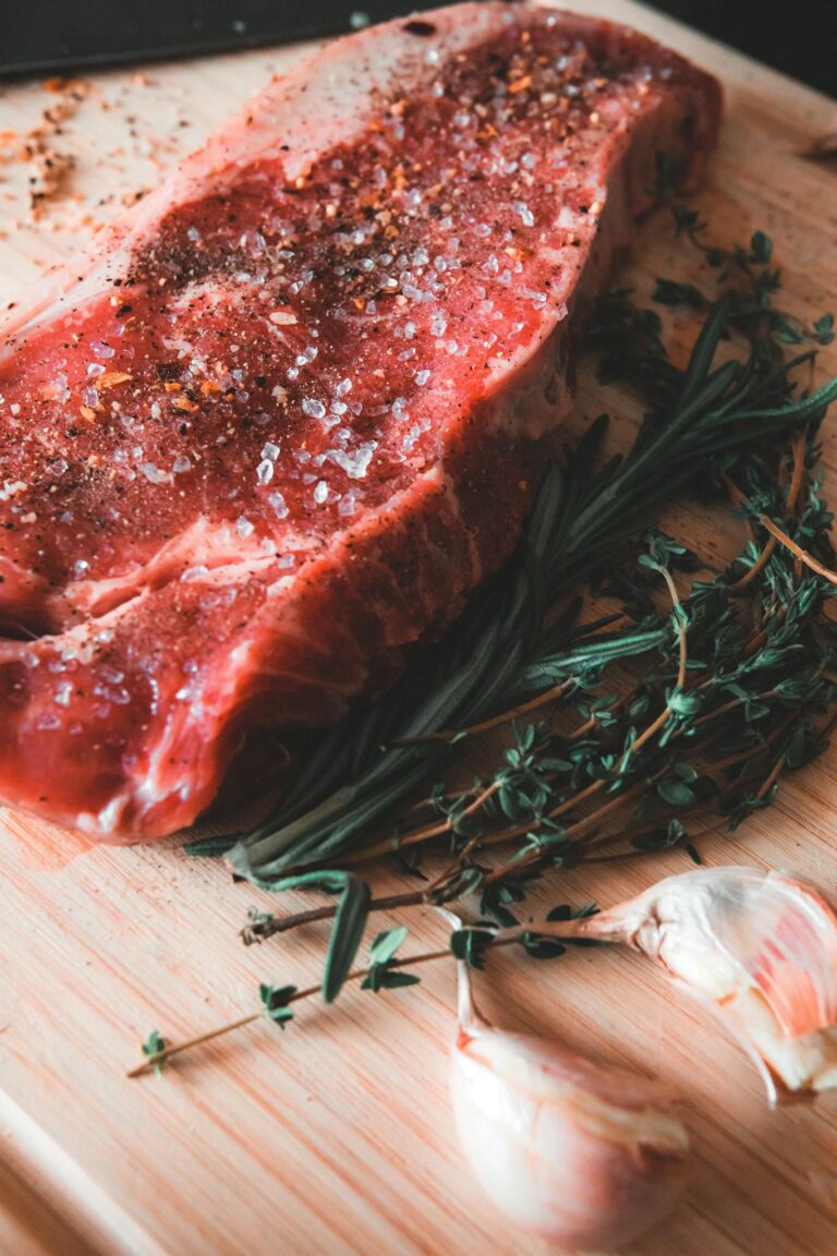 A raw beef steak seasoned with salt and spices, surrounded by fresh rosemary, thyme, and garlic on a cutting board.