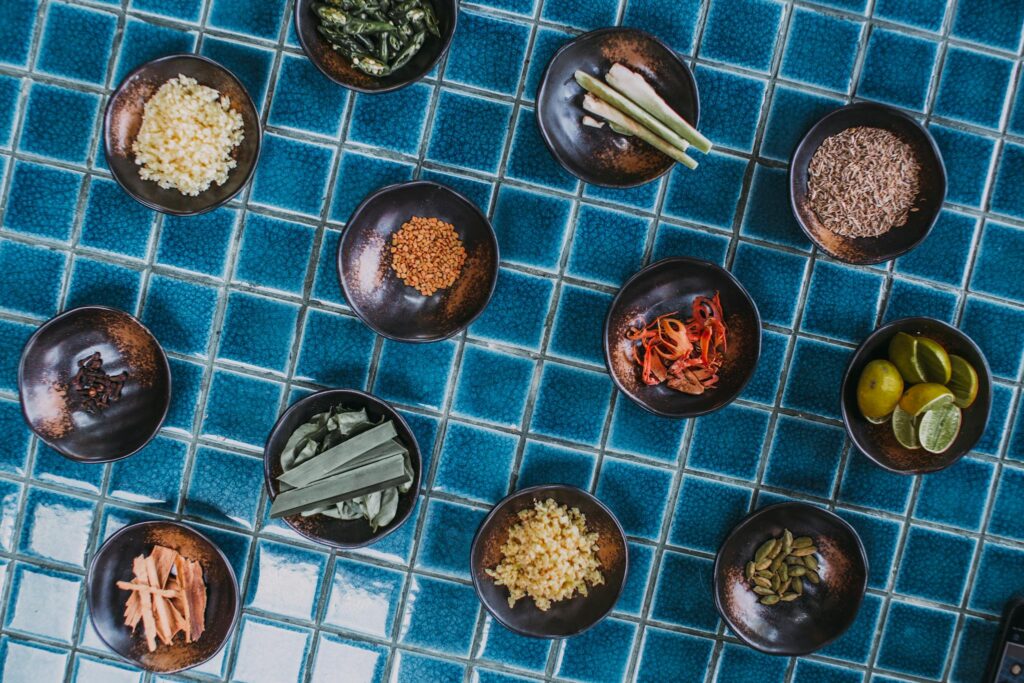 Assorted spices and ingredients in bowls arranged on blue tiles, top-down view.