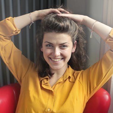 Cheerful woman sitting indoors, smiling brightly with hands behind head, wearing a yellow shirt.